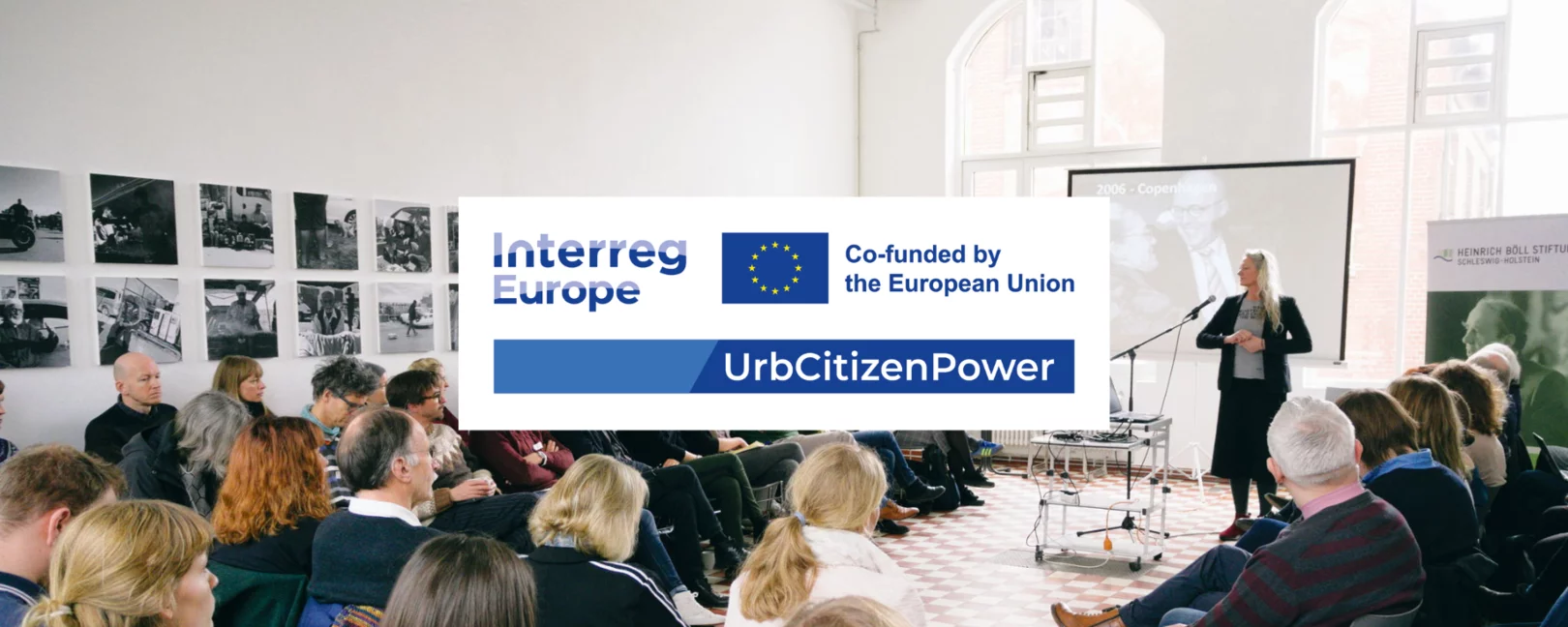 Im Hintergrund sind sitzende Menschen zu sehen, die einer stehenden, einen Vortrag haltenden Person, zuhören. Mittig im Vordergrund ist das Logo von UrbCitizenPower. Auf diesem befinden sich in Blau geschrieben die Schriftzüge 