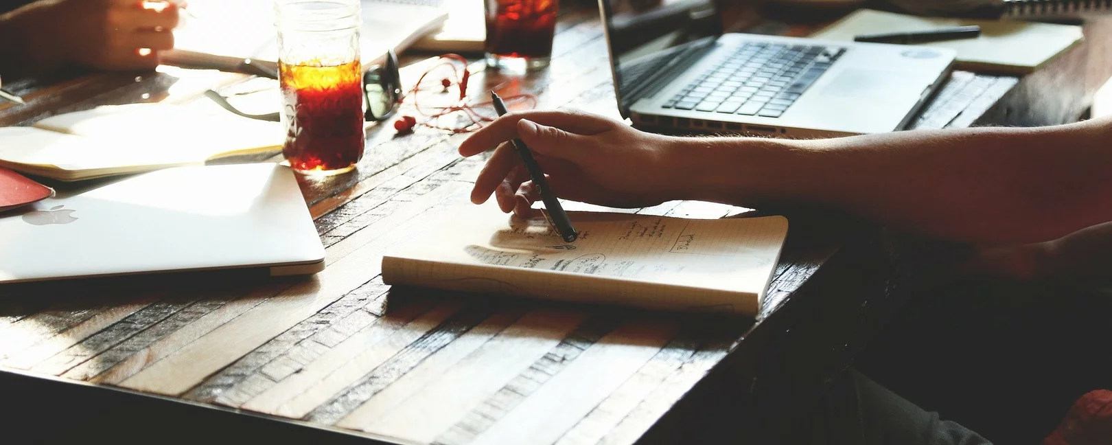 Nahaufnahme von einem Tisch. Auf dem Tisch liegen Blöcke, stehen Gläser und ein Laptop. Man sieht die Arme und Hände von Leuten, die Stifte in den Händen halten.