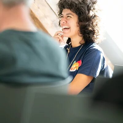 Eine Person lacht und sitzt auf einem Stuhl. Sie scheint im Gespräch mit anderen zu sein.