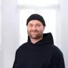 Lasse Trede Potrait von Lasse Trede. Ein junger Mann mit dunklem Dreitagebart lächelt in die Kamera. Er trägt einen schwarzen Hoodie und eine schwarze Mütze.