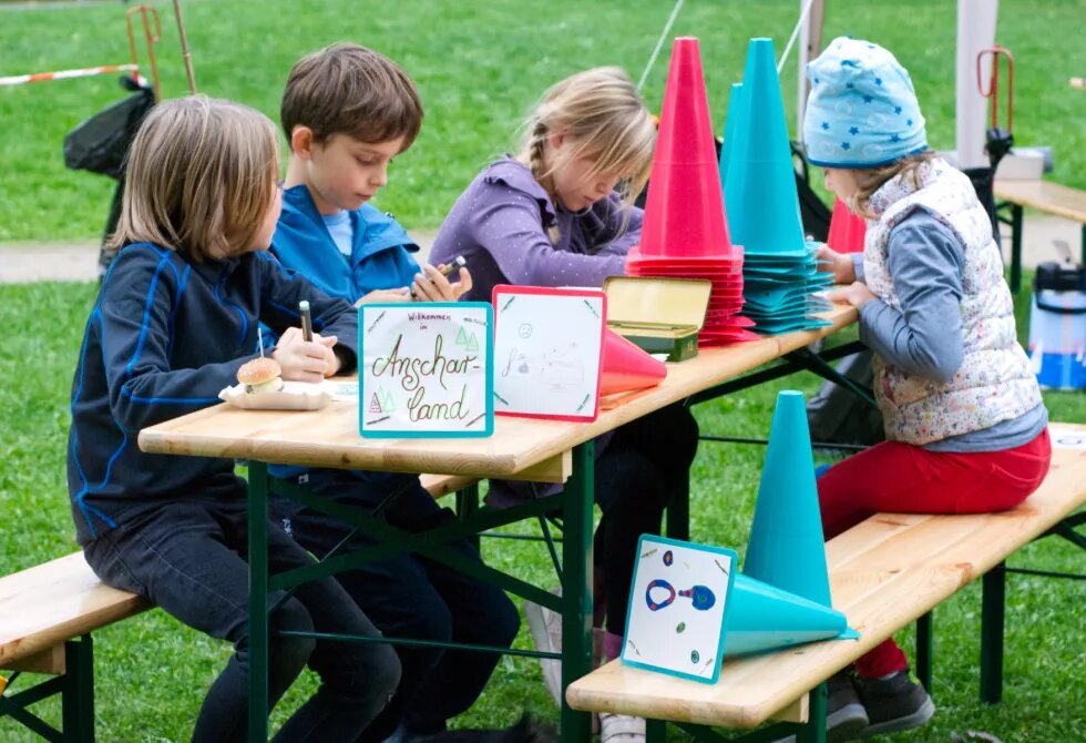 Zwei Mädchen und zwei Jungen sitzen an einer Bierzeltgarnitur im Park und malen. Auf dem Tisch stehen gestapelte bunte Kegel, einige davon liegen umgekippt auf der Seite und zeigen ein Bild auf der Unterseite. Unter dem Tisch liegt ein Hund.