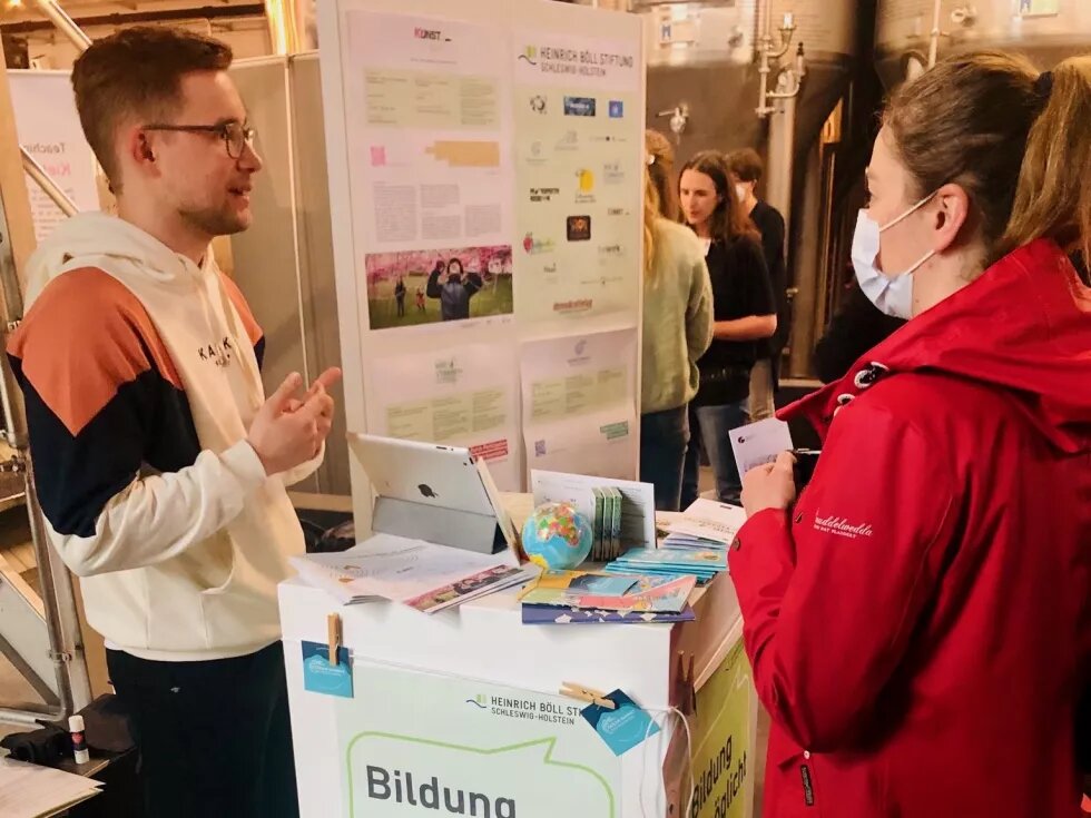 Austausch bei der BNE-Konferenz Links steht ein junger Mann, rechts eine junge Frau. Zwischen ihnen ist ein Pult mit Flyern und Plakaten aufgestellt. Der Mann spricht mit edr Frau. Im Hintergrund sieht man eine Plakatwand der Heinrich-BöllStiftung Schleswig-Holstein.