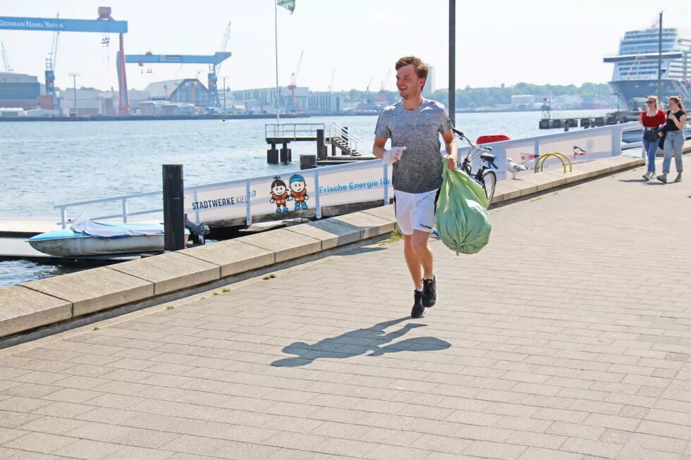 Ocean Summit Plogging Ein jüngerer Mann joggt an einer Promenade entlang. in der linken Hand hält er einen grünen Müllbeutel. Im Hintergrund sind Wasser, ein Kreuzfahrtschiff und Kräne zu sehen.