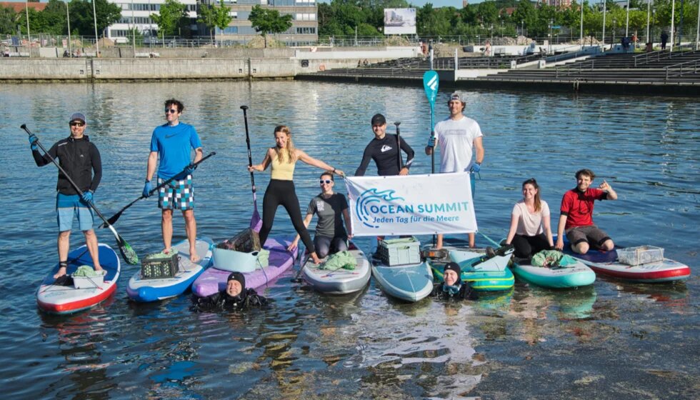 Ocean Summit SUPs Auf dem Wasser liegen mehrere Stans-Up-Paddel-Boards nebeneinander. Auf den Boards stehen Leute mit Paddeln in der Hand. Zwei von ihnen halten ein Banner auf dem steht: Ocean Summit - jedena Tag für die Meere.