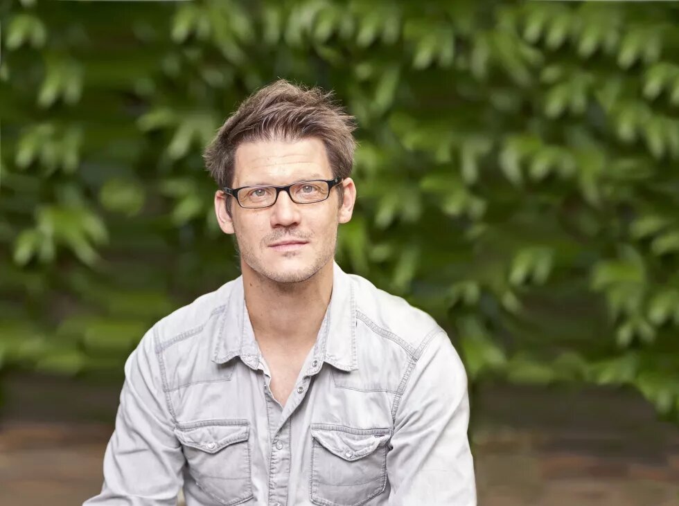 Torsten Schäfer Porträt von Torsten Schäfer. Ein mittelalter Mann sitzt vor einem grünen Strauch. Er trägt ein graues Jeanshemd und eine Brille. Er hat kurze braune Haare.