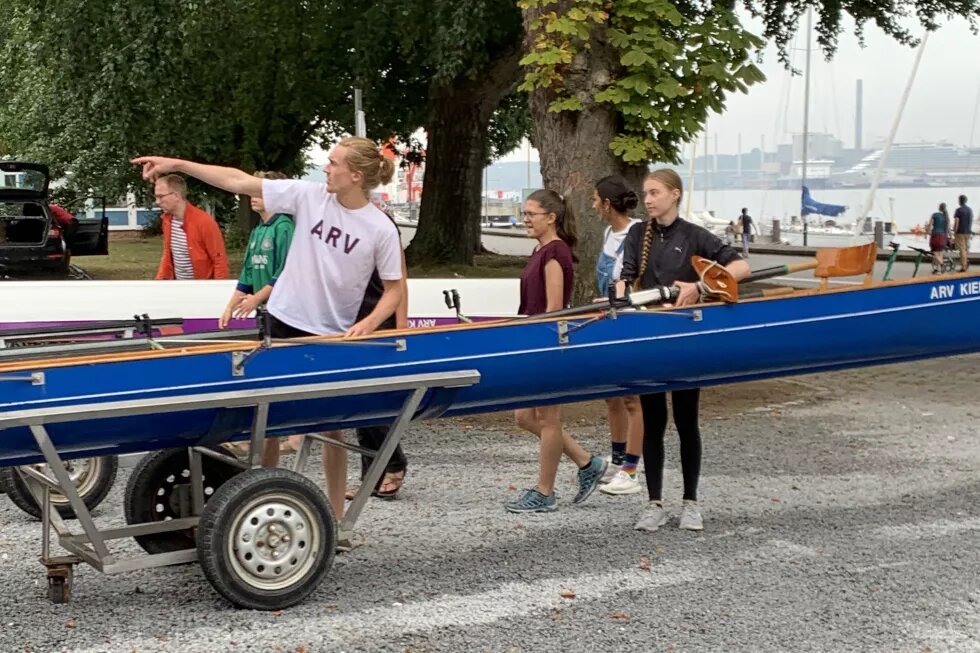 Rudern beim Sommerfest Mehrere Personen stehen um ein Ruderboot herum. Eine Person deutet nach links. Im Hintergrund sieht man Wasser und Hafen.