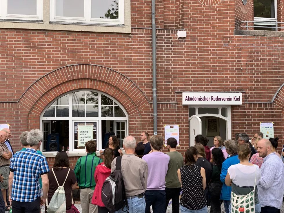 Vor einem Backsteingebäude stehen Uta Röpcke und Malte Krüger und halten ein Grußtwort. Vor ihnen steht mit dem Rücken zur Kamera eine große Gruppe Menschen.