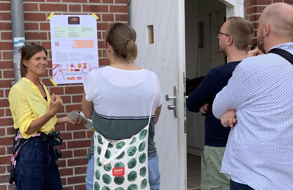 Andrea Cederquist steht vor einem Plakat der Salons des guten Lebens und spricht mit drei Leuten.