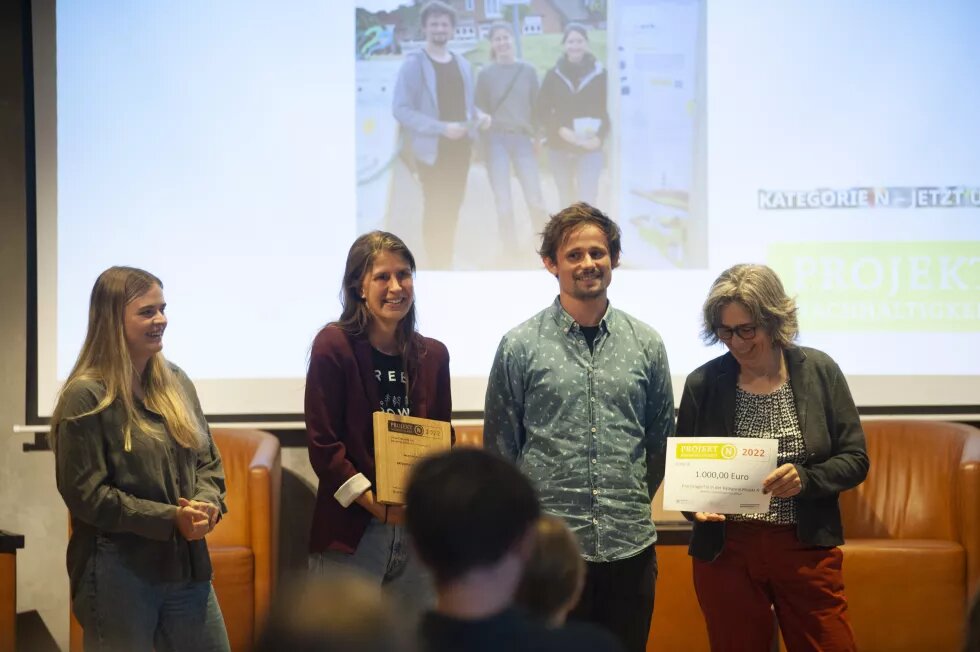 Preisverleihung bewirk Vorne stehen viere erwachnsene Personen, die eine Urkunde in der Hand halten. Im Hintergrund ist ein Foto an eine Wand projiziert.