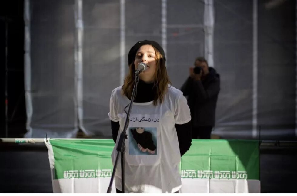 Esther Mischke Esther Mischke trägt eine schwarze Kappe und ein bedrucktes T-Shirt. Sie spricht in ein Mikrophon. Hinter ihr sind Teile der iranischen Flagge zu sehen.