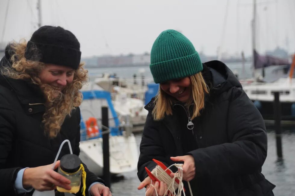 Zwei Personen (Stefanie Sudhaus links, Louisa Dellert links) halten eine Schnur und ein Gefäß in den Händen. Im Hintergrund ist ein Hafen.