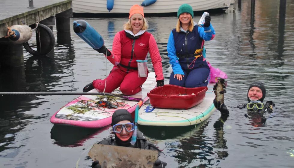 Zwei Personen sitzen in dicken Anzügen auf zwei SUPs und halten Gegenstände in den Händen. Im Wasser sind zwei Personen in Tauchanzügen.