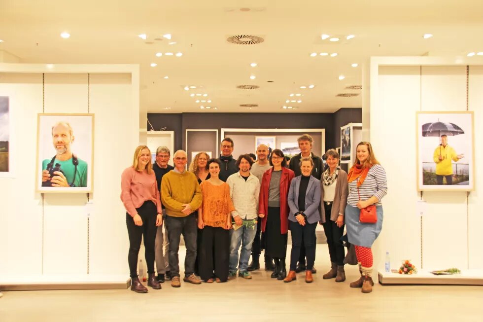 Vernissage Meeresmenschen Eine Gruppe von Menschen steht in einem Raum im Nordlicht Einkaufszentrum in Kiel und schaut in die Kamera. Um sie herum hängen Bilder der Ausstellung Meeresmenschen.