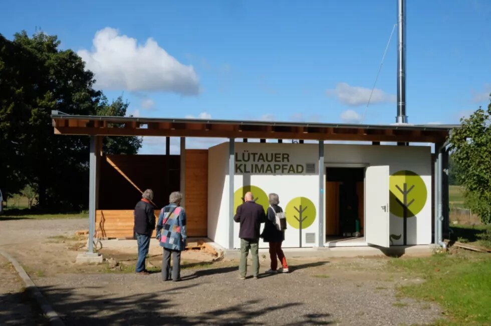 Lütauer Klimapfad Auf einem mit Bäumen bemalten Gebäude in Lütau steht Lütauer Klimapfad. Davor stehen 4 Personen, die sich das Gebäude anschauen.