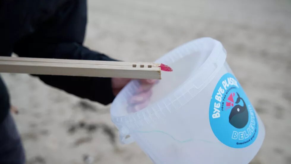 frutti di mare Auf einem Eimer steht "Bye bye Plastik" Mit eine Holzzange tut jemand Müll vom Strand in den Eimer.