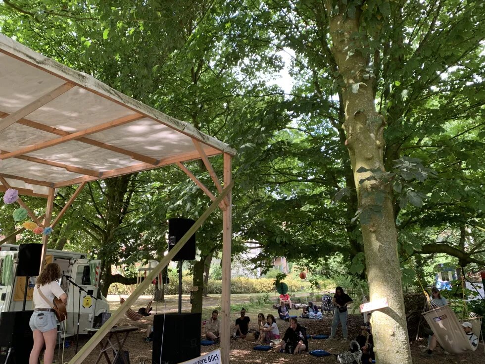 versnuven: ALTE MU x Böll SH Auf einer Bühne spielt Musikerin Lucie Glang im Rahmen von versnuven: ALTE MU x Böll SH Gitarre. Die Bühne befindet sich im Vorgarten des ALTE MU Impuls-Werks e.V. Unter Bäumen sitzt PPublikum, das der Musik lauscht.