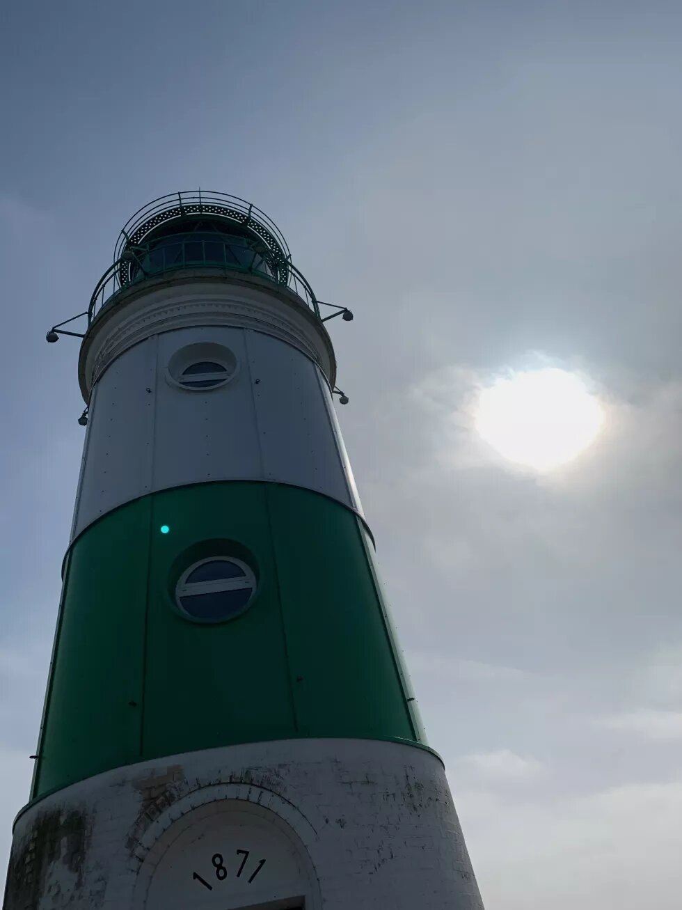 Der grün-weiße Leuchtturm von Schleimünde von unten fotografiert. Rechts ist die Sonne vor blauem Himmel.