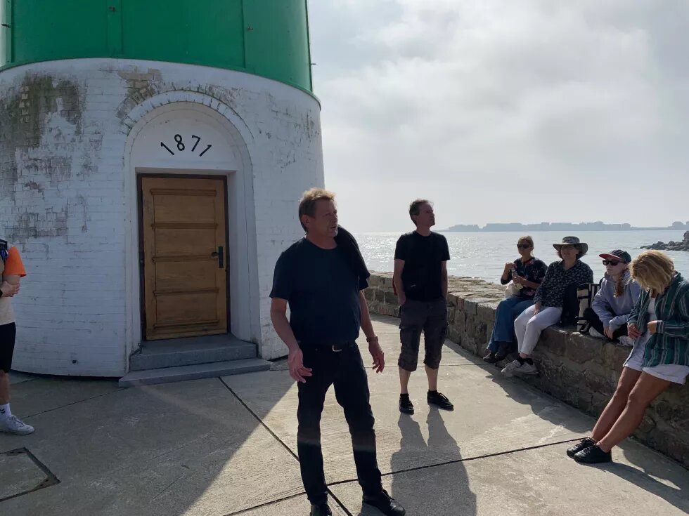 Jens von der Lighthouse Foundation steht vor dem Schleimünder Leuchtturm und erklärt. Rechts auf der Kaimauer sitzen Mitarbeitende der Böll SH. Im Hintergrund ist die Ostsee.