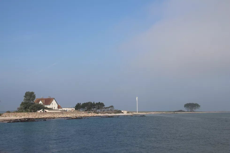 Vorne ist die Ostsee. Hinten steht das weiße Lotsenhaus auf der Insel Schleimünde. Darum sind Bäume und Strand.