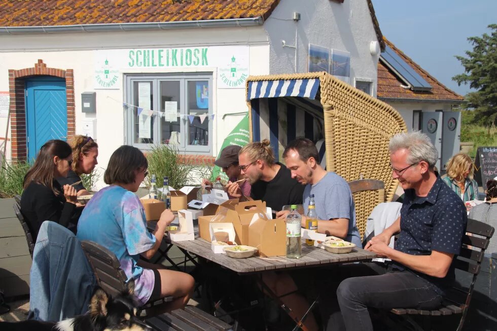 Sechs Mitarbeitende der Böll SH sitzen um einen Tisch, auf dem Lunchpakete stehen, und essen. Im Hintergrund ist ein weißes Haus.