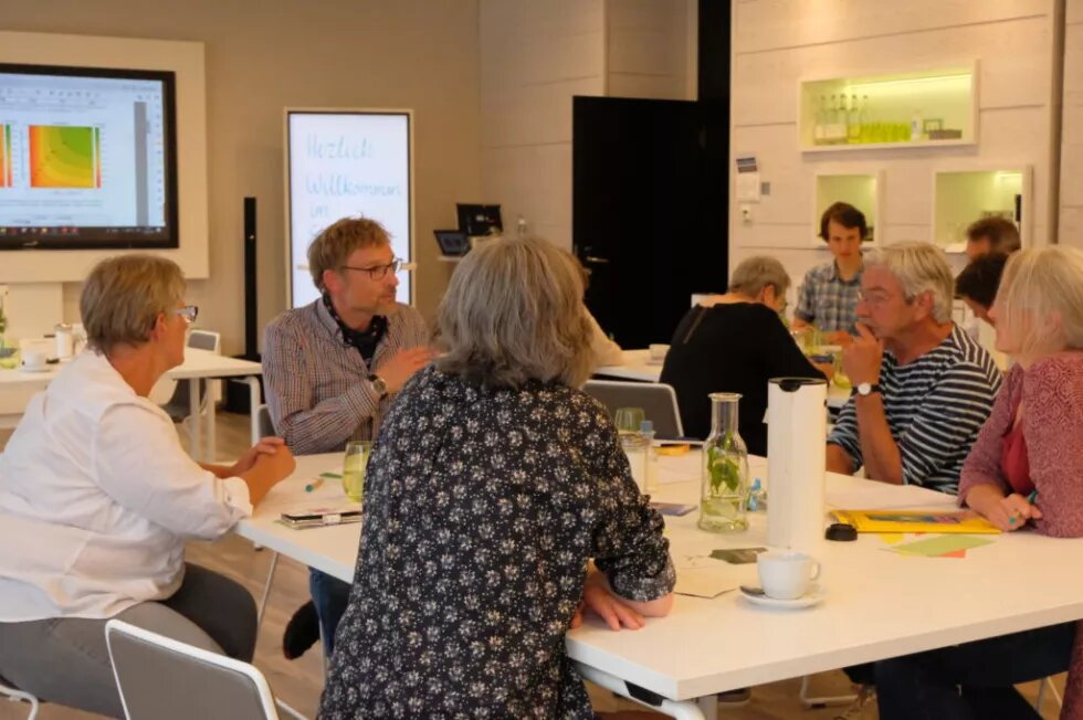 bewirk – gemeinsam fürs Klima Bei einer Energiewende-Ideensession von bewirk sitzen mehrere Personen um einen Tisch herum. Im Hintergrund leuchtet ein Bildschirm mit bunten Grafiken.