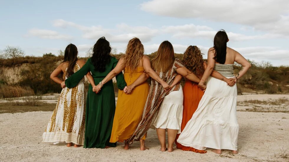 Sechs Frauen stehen nebeneinander im Arm mit dem Rücken zur Kamera zusammen. Sie halten ihre Hände hinter dem Rücken der jeweils neben ihnen stehenden Frau. Alle tragen Kleider. Im Hintergrund sind Dünen zu erkennen.