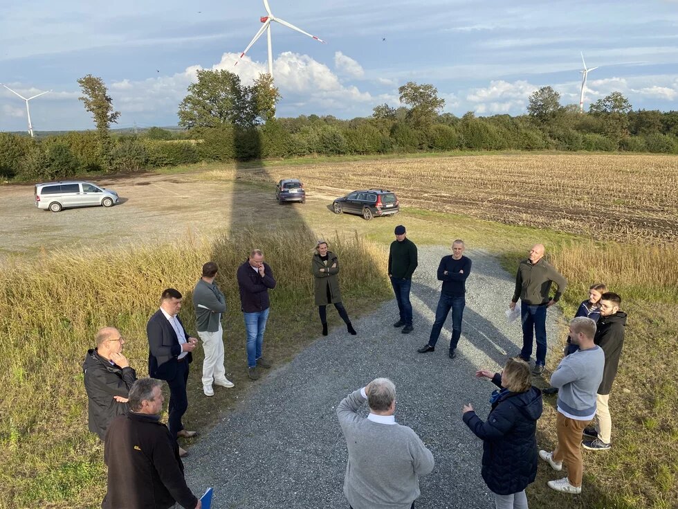 Eine Gruppe von Menschen steht im Halbkreis auf einem Kiesweg in einer ländlichen Umgebung. Im Hintergrund sind Windkraftanlagen und ein abgeerntetes Feld zu sehen. Einige Autos sind auf einem unbefestigten Parkplatz abgestellt. Die Personen scheinen sich im Gespräch zu befinden, während sie sich die Umgebung ansehen.