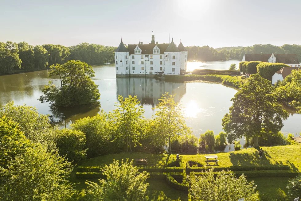Das Glücksburger Wasserschloss liegt in einem See. Die Sonne scheint über den Türmen. Drumherum ist Wald.