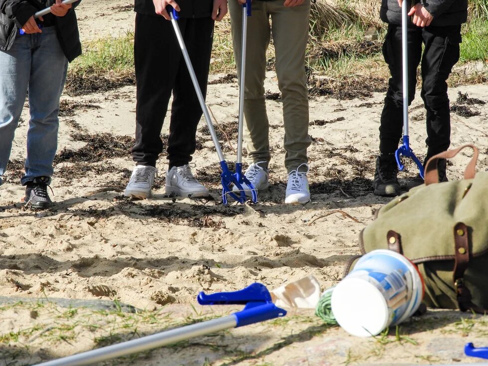 Vier Jugendliche sammeln Müll am Strand mit Müllzangen.