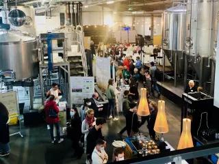 BNE-Konferenz in der Lille Brauerei Man sihet eine große Halle, in der Braukessel für Bier stehen. Zwischen den KEsseln stehen Stände und Plakate, an denen viele Menschen stehen. Außerdem gibt es ein Buffet.