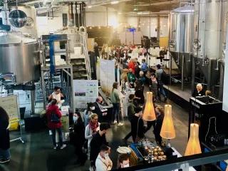 Man sihet eine große Halle, in der Braukessel für Bier stehen. Zwischen den KEsseln stehen Stände und Plakate, an denen viele Menschen stehen. Außerdem gibt es ein Buffet.