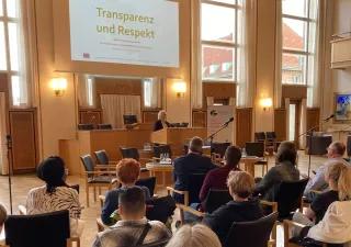 Man sieht sitzendes Publikum von hinten. Vorne steht am Podium eine blonde Person. Dahinter ist Transparenz und Respekt an die Wand projiziert.