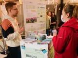 Austausch bei der BNE-Konferenz Links steht ein junger Mann, rechts eine junge Frau. Zwischen ihnen ist ein Pult mit Flyern und Plakaten aufgestellt. Der Mann spricht mit edr Frau. Im Hintergrund sieht man eine Plakatwand der Heinrich-BöllStiftung Schleswig-Holstein.